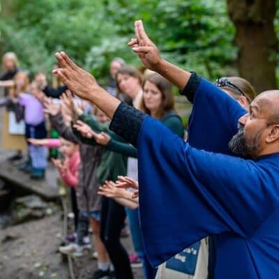 A dancer in a blue robe, hands raised, with a group of people copying his movements in an outdoor setting with trees behind