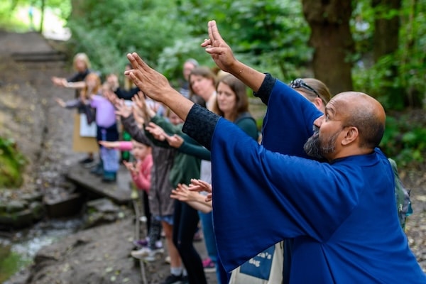A dancer in a blue robe, hands raised, with a group of people copying his movements in an outdoor setting with trees behind